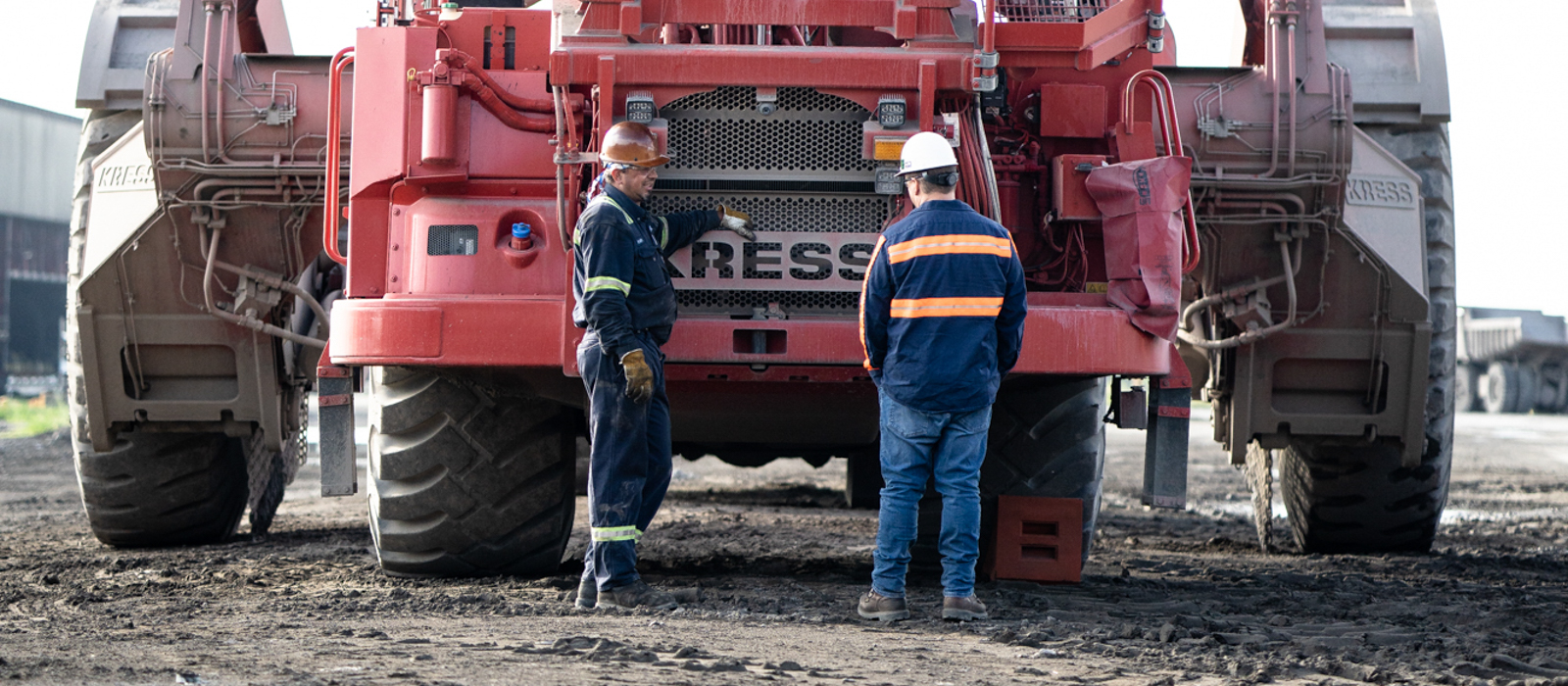 Two Employees Wearing Hard Hats And Neon Safety Vests Standing In Front Of Red Kress Heavy Equipment At Project Site