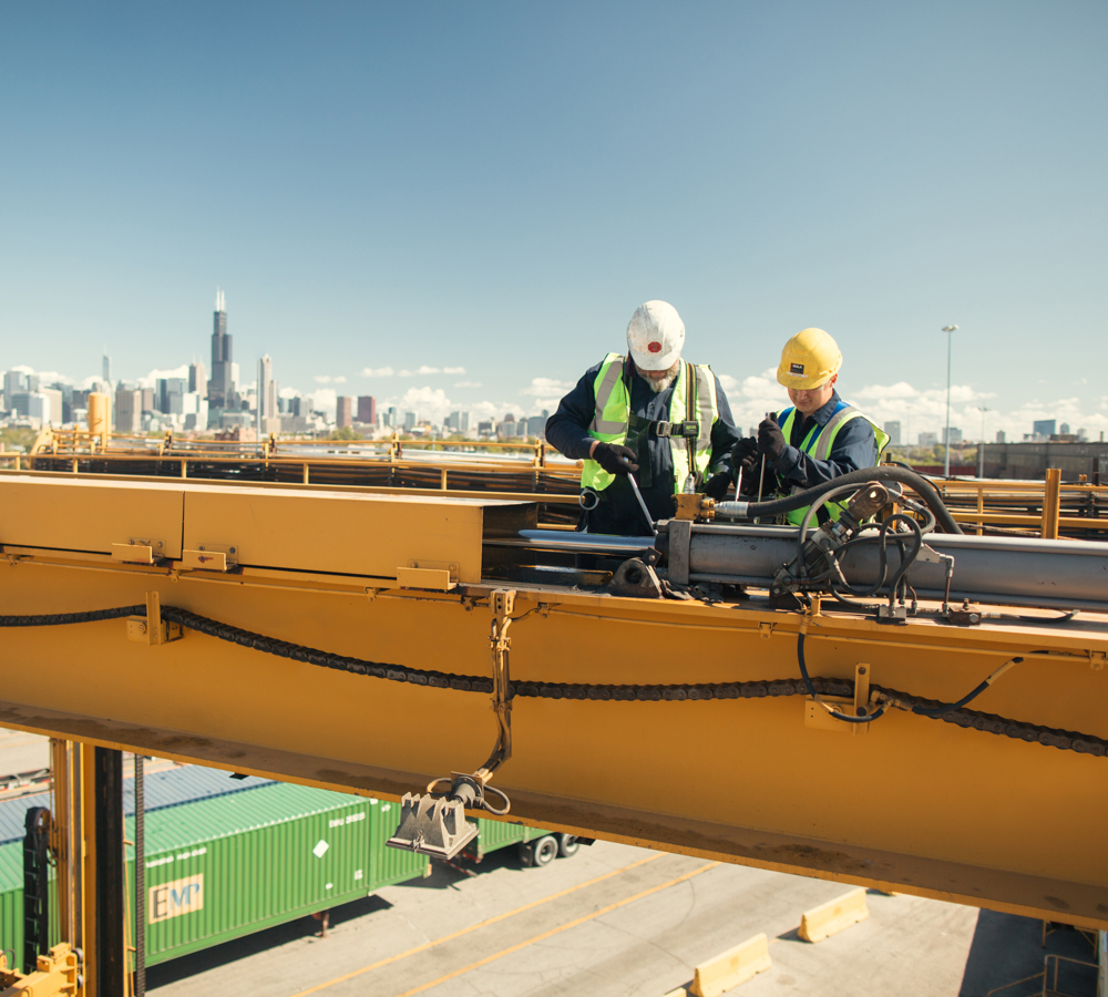 Two Employees Wearing Hard Hats And Neon Safety Vests Working On Industrial Equipment