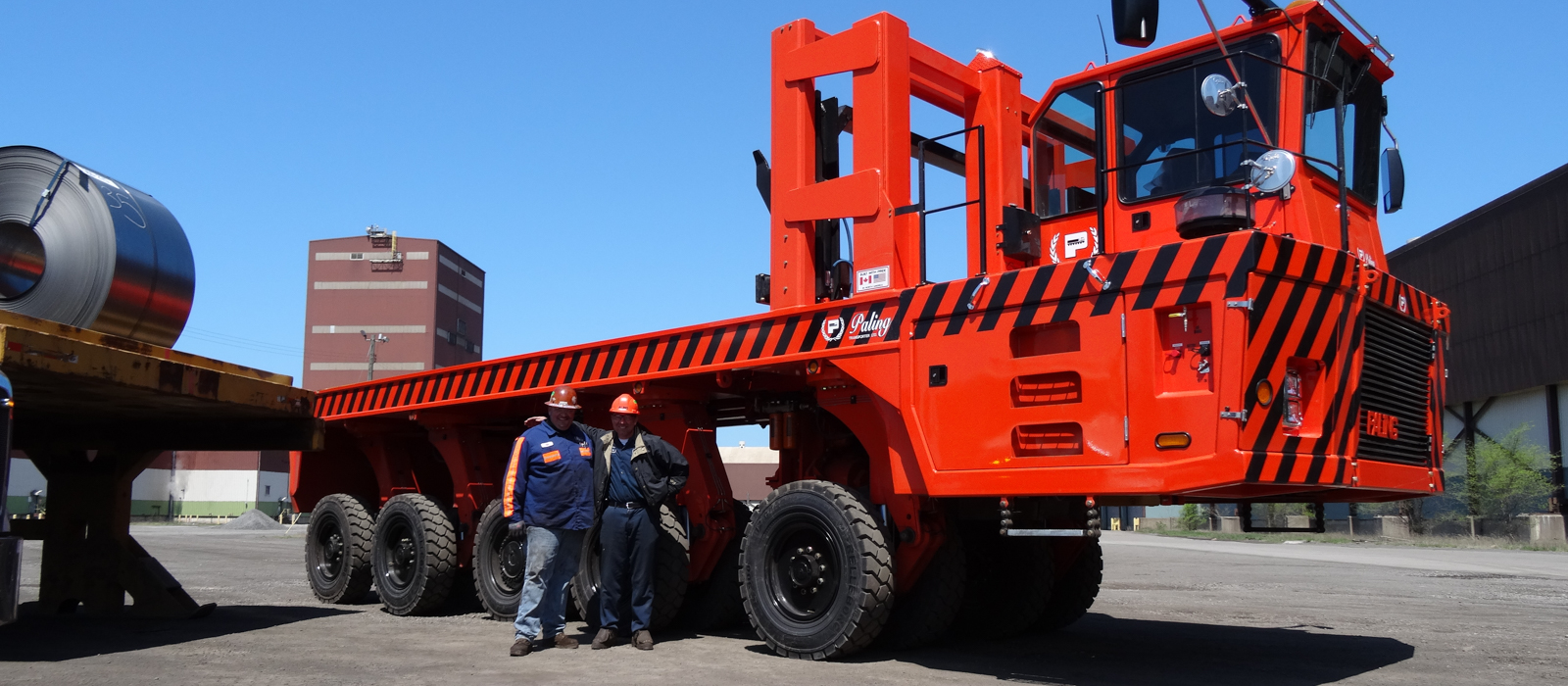 Two Bulk Equipment Corp. Employees Wearing Hard Hats Standing In Front Of A Bright Red Transporter