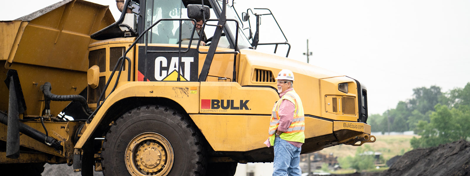 Bulk Equipment Employee Wearing Hard Hat, Neon Vest & Safety Glasses Next To Cat® Heavy Equipment At A Landfill Job Site